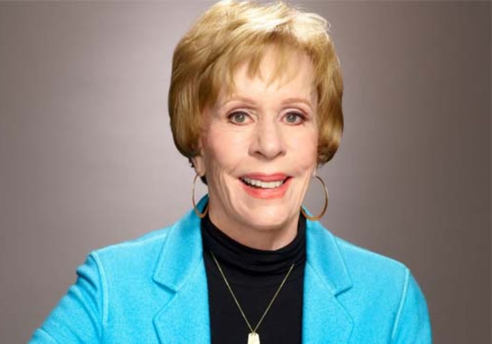 A photo of Carol Burnett smiling and holding a microphone, dressed in a red blazer with a black blouse and pants.